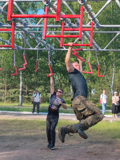 Two men are participating in an obstacle course on a sunny day in Sertolovo, Russia. In the foreground, a man in a black t-shirt and camouflage pants is swinging from a red monkey bar, his legs extended. Behind him, another man in a dark t-shirt and dark pants is reaching for a similar red bar. Both men appear to be in the middle of the course, with other obstacles visible above them. In the background, several spectators are observing the event. The terrain is a mix of dirt and grass, with trees and a blue sky overhead. The man in the foreground is wearing a bib with the number "22608".