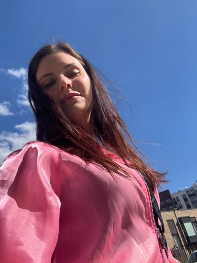 A low-angle shot captures a young woman looking upwards against a bright blue sky with scattered white clouds. She has long, dark brown hair that is slightly windswept, framing her face which is turned towards the camera. She is wearing a bright pink, shiny bomber jacket with the zipper partially open. In the background, the upper floors of modern apartment buildings and a street lamp are visible under the clear sky. The scene appears to be outdoors in an urban environment. The provided hint of the city of Novoselye, Russia, is a geographical location.