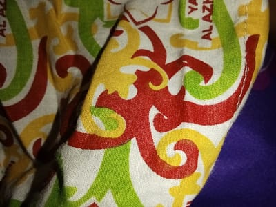 A close-up forensic image depicts a section of a patterned textile, likely fabric, exhibiting folds and creases. The primary design, set against a white background, features interlocking abstract curvilinear motifs in red, yellow, and green. Visible white text elements embedded within the pattern include fragmented characters forming "YASAN" and "AL AZH", referencing "Yayasan Al Azhar". The fabric texture is discernible, showing a fine weave. A dark purple, out-of-focus surface is visible in the background on the right. No individuals are present. This scene is situated in Ciputri, Indonesia.