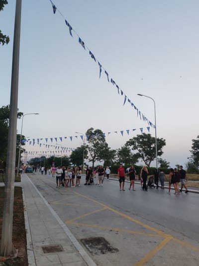 The image depicts a street scene in Alexandroupolis, Greece, likely during an outdoor event or festival. A street is lined with people walking and gathered in groups. The sky is clear and a pale blue, suggesting it is either early morning or late evening. A prominent feature is strings of blue and white triangular flags, resembling the Greek flag, draped across the street from lampposts. Trees line the sides of the street, and lampposts are visible. There is a sidewalk on the left with a few people walking on it. On the street, there are cars parked and driving, and some people are pushing strollers. The overall atmosphere appears festive and public.