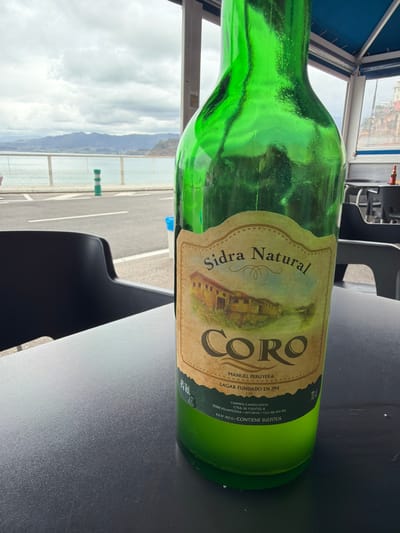 The scene depicts a green glass bottle of "Sidra Natural Coro" cider, positioned on a dark, textured tabletop in a semi-enclosed establishment. The bottle's label is prominent, featuring an illustration of a building and text including "Manuel Peruyera," "LAGAR FUNDADO EN 1914," and "33300 VILLAVICIOSA - ASTURIAS." The bottle is largely full with a light green liquid. To the left of the bottle, a black chair is partially visible.

Beyond the table, a paved road or promenade stretches alongside a body of water, separated by a light-colored railing. A green and white striped bollard stands on the road. In the distance, a range of mountains is visible under a cloudy sky. To the far right, through a window or opening, a cliffside adorned with buildings is apparent, along with a distant table holding a small red object. The overhead structure of the establishment includes a blue canopy. The environment is coastal, indicative of a location such as Colunga, Spain.
