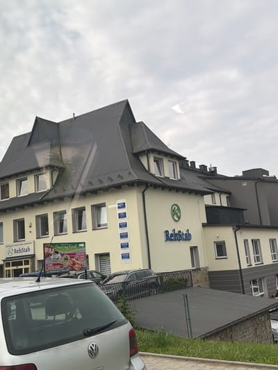The image shows a light yellow building with a dark grey, multi-pitched roof. A large, dark grey sign with the text "RehStab" and a green circular logo is visible on the left side of the building. To the right of this sign, a green and white billboard advertises a shop called "Sklepik Smaczny Kacik", featuring images of food and drinks. The building has multiple windows, some with white frames and others with dark frames. Several cars are parked in front of and around the building, with a silver car prominently in the foreground, partially obscuring the lower part of the building. The environment appears to be an urban setting with a cloudy sky. Based on the signs, the building is located in Limanowa, Poland.