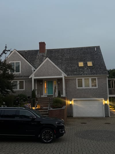 The image shows a two-story house with gray cedar shingle siding and a gray cedar shingle roof in Provincetown, United States of America. A red brick chimney rises from the roof. The house has several windows, including two dormer windows on the upper level of the roof and three rectangular windows on the second floor above a white garage door. A light blue front door is visible on the porch, accessible by a set of brick steps. A black SUV is parked in the driveway, which is paved with cobblestones. The scene appears to be set in the evening, with lights on in the house and exterior light fixtures illuminating the driveway and porch. Lush green landscaping with pink and white flowers surrounds the front of the house.