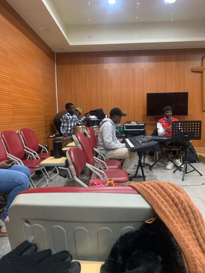 This image depicts an indoor musical practice or rehearsal session occurring in a room located in Shenyang City, China. The room features light-colored flooring, wood-paneled walls, and a white suspended ceiling with recessed lighting. A dark, wooden cross is mounted on the right wall, above a wall-mounted flat-screen television. Small, colorful decorative items are suspended from the ceiling.

Five individuals are visible. On the left, partially obscured, a person in a plaid shirt is positioned near a drum kit, appearing to engage with the drums. To their right, a person wearing a light grey hoodie and a black baseball cap is seated, operating an electronic keyboard placed on a stand. Further to the right, a person dressed in a red and white varsity-style jacket and a black beanie is seated at another electronic keyboard, positioned behind a black music stand with an attached microphone. This individual appears to be either playing the keyboard or singing. Audio equipment, including amplifiers and mixers, is visible on a rack between the two keyboardists. Several red-backed chairs with metal frames, some featuring foldable writing tablets, are arranged in rows. In the immediate foreground, a light-colored portable cooler or container is visible, topped with an orange knitted scarf. Black gloves are partially visible on a surface in the bottom left foreground. A black travel mug rests on a side table attached to one of the red chairs, and a red tambourine with yellow accents is on another chair.