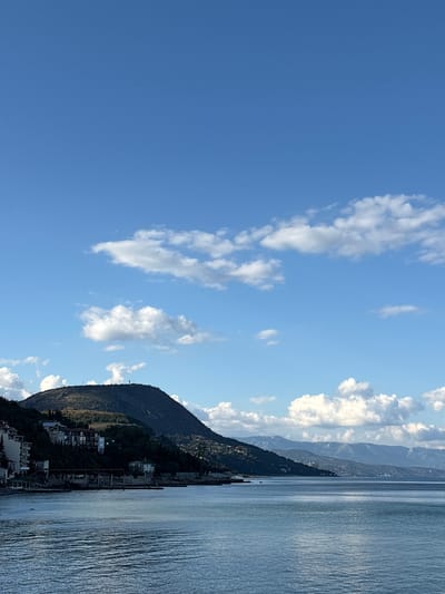 The image depicts a coastal scene with a body of water in the foreground. In the background, a large, dark, tree-covered mountain dominates the landscape. Along the slopes of the mountain, several buildings are visible, suggesting a settlement. The sky is blue with scattered white clouds. The water appears calm with gentle ripples. The overall impression is of a scenic, possibly residential, coastal area. The location is identified as the city of Utios, Ukraine.