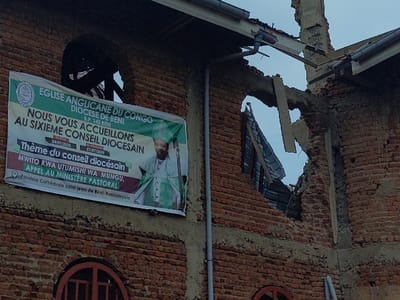 An outdoor, eye-level photograph depicts a damaged brick building, identified by a banner as the Saint John Cathedral Parish of Beni Kasabinyole, Anglican Diocese of Congo, located in Beni, Democratic Republic of the Congo. The building's upper facade exhibits extensive structural damage, characterized by large, irregular breaches in the reddish-brown brickwork. Twisted metal roofing sheets and broken wooden beams protrude from these openings, through which a cloudy sky is visible. Grey downspouts are affixed vertically to the brick wall. A large, predominantly green and white banner hangs on the left side of the facade, announcing the "Sixième Conseil Diocésain" scheduled from "24/07/2035" to "27/07/2035," and displaying an image of a man in green and white clerical vestments. Arched window openings are present; the upper left window appears open, and the lower right window contains a decorative metallic insert. No individuals are visible in the scene.