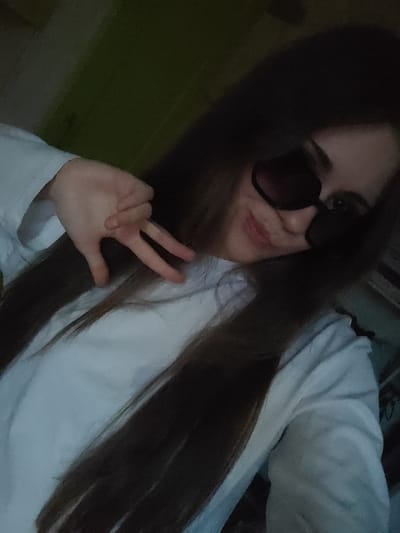 A young woman with long, dark hair and dark sunglasses is taking a selfie. She is wearing a white long-sleeved shirt. Her left hand is raised, with her index and middle fingers extended, forming a peace sign. She is smiling at the camera. The background is dark and blurry, with a hint of green on the left side. The lighting is dim. The image was likely taken indoors, in a private residence in Svietlahorsk, Belarus, as indicated by the background which appears to be a wall in a room.
