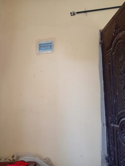 The image depicts an interior wall with a pale yellow hue. Embedded in the wall, slightly to the left of the center, is an electrical panel with a light blue cover. The panel is rectangular and appears to be a standard household electrical box. To the right of the panel, a portion of a dark brown, ornately carved door is visible, suggesting it is either an entrance or an internal doorway. Above the electrical panel, a dark rod is mounted horizontally on the wall, with a small, decorative metal piece attached to its left end. There is also a hook hanging from the rod. The lower left corner of the image shows a dark surface, possibly furniture, with some crumpled fabric, including a red item and lighter colored cloth, draped over it. The environment appears to be a room in a building, possibly residential, located in Gulak, Nigeria.