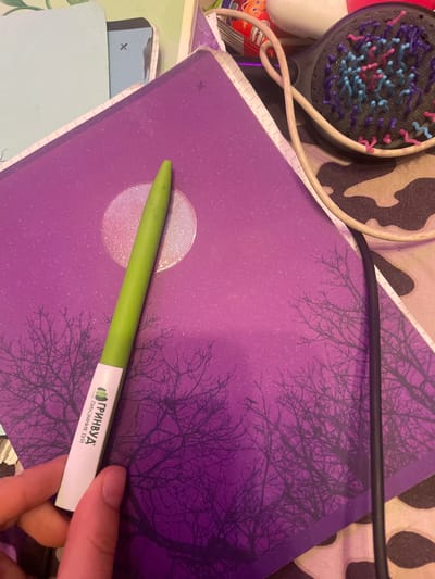A close-up shot shows a person's hand holding a green pen over a purple notebook with a silhouette of trees and a full moon. The notebook is lying on a patterned surface, possibly fabric. To the right of the notebook, a black cable and a beige wire are visible, and behind that, a black hairbrush with multi-colored bristles is partially in view. The scene is indoors. There is no indication of Romanovka, Russia in the image.