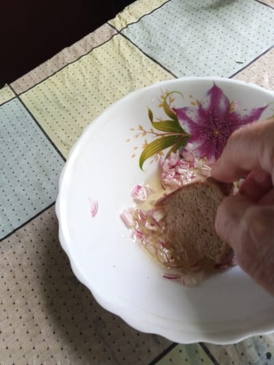 A hand is dipping a piece of bread into a white bowl. The bowl contains a liquid, chopped red onions, and a floral design featuring a large pink and purple flower with green leaves. The bread appears to be whole grain and is partially submerged in the liquid. The scene is set on a table covered with a patterned tablecloth, featuring squares of yellow, white, light blue, and beige, with small black dots. The tablecloth has a woven texture. The lighting suggests it is daytime. The overall impression is a home setting, likely in Luka, Ukraine, where someone is preparing or consuming a simple dish.