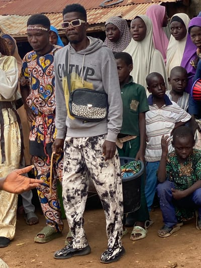 In the city of Rigachikun, Nigeria, a group of people are gathered outdoors, in front of a structure with a corrugated metal roof. The ground is composed of dry, brown soil.
The primary subject is a man standing prominently in the foreground, wearing a grey hooded sweatshirt and pants with a black and white marble pattern. He has a crossbody bag with a leopard-print front pocket. He wears sunglasses and a headband adorned with shells. He holds a thin wooden stick.
To his left, a man in a vibrantly patterned traditional outfit, featuring blue, yellow, and red motifs, stands with a similar headband. To the right of the primary subject, a group of children are present. One child, wearing a green patterned shirt and blue pants, is seated on the ground. Other children stand around him, some wearing t-shirts and others traditional attire. Several women, some wearing hijabs in various colors including purple, light pink, and cream, are visible in the background. The overall scene appears to be a social gathering or community event.