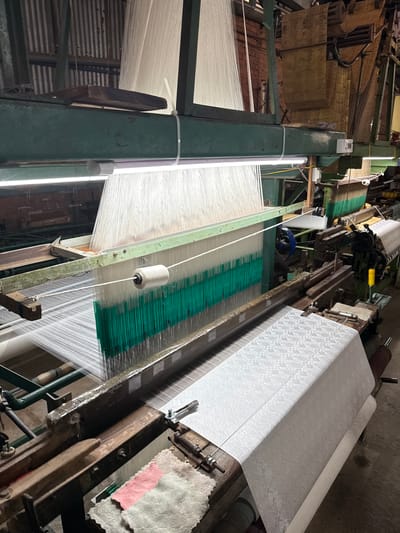The image depicts a loom in operation within a textile factory in Nam Ban, Vietnam. The loom is in the process of weaving a white fabric with an intricate pattern, which is visible on the large roll of finished fabric to the right. Thousands of white threads are suspended vertically, forming the warp of the fabric. A single white spool of thread is mounted on the loom, ready to be woven into the fabric. The loom itself is constructed from a combination of green-painted metal and weathered wood. In the background, corrugated metal walls and brickwork suggest the industrial setting. To the left of the loom, stacks of what appear to be punched cards, used for programming the loom's pattern, are visible. The lighting appears to be from overhead fluorescent tubes. The overall scene is one of an active manufacturing process.