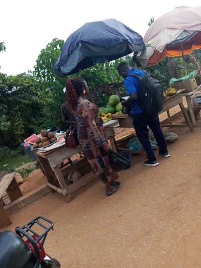 In Galadimawa Village, Nigeria, a scene unfolds at a roadside market. Two individuals are interacting near makeshift wooden stalls. A woman, adorned in a patterned dress and a maroon handbag slung over her shoulder, stands facing away from the camera. She has auburn hair styled in braids. Beside her, a man wearing a blue t-shirt and dark pants, carrying a black backpack, faces towards the stalls. He appears to be looking at items on display.
The market stalls are laden with produce. One stall displays several coconuts and sliced watermelons, arranged on a white cloth. Another stall, further back, shows bunches of bananas and what appear to be gourds or large fruit. Two large umbrellas, one blue and one with orange and white fabric, are set up to provide shade over the stalls. The ground is a dusty, reddish-orange earth, characteristic of the Nigerian landscape. In the foreground, the rear of a red motorcycle is visible, with a black seat and rack. The surrounding environment is lush with green foliage, indicating a rural or semi-rural setting within the village. The overall impression is of a bustling, informal market scene.