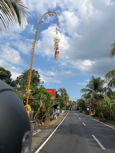 The image shows a street scene in Selemadeg Barat, Indonesia, on a partly cloudy day. A tall, yellow, arched decoration with a hanging ornament is prominently featured in the sky on the left side of the road. The road itself is paved with asphalt and has white dashed lines. On either side of the road, there are lush green trees, palm trees, and some buildings. Some of the buildings appear to be small shops or stalls. There are also some red and white flags visible along the roadside. The foreground on the left is dominated by the dark, curved surface of what appears to be a helmet, suggesting the photo was taken from a motorcycle or scooter. The overall impression is that of a rural or semi-rural street decorated for a local celebration or festival.