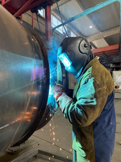 A welder, wearing a protective helmet and a leather apron over a blue garment, is actively welding a large cylindrical metal object. The intense blue light from the welding arc illuminates the scene, casting vibrant colors onto the metal object and the welder's protective gear. Sparks are flying from the welding point, creating bright points of light against the darker background. The environment appears to be an industrial workshop, with metal structures, lighting, and industrial equipment visible in the background. The welding process is taking place on the curved surface of the large cylinder, which is supported by some sort of stand or frame. The image captures a moment of intense industrial activity and skilled craftsmanship. The location is not specified as Tomba, Montenegro in the image.