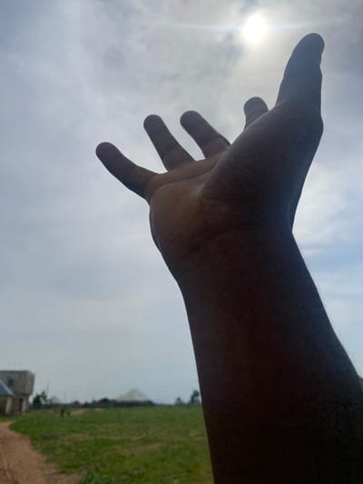 The media file shows a close-up shot of a person's dark-skinned hand reaching out, palm facing forward, against a bright, cloudy sky. The sun is visible as a bright, slightly diffused orb in the upper left portion of the frame, suggesting it is daytime, likely mid-morning or late afternoon given the softness of the light. The hand is in focus, with the fingers slightly spread and the thumb extended. The arm extends out of the frame to the right.

In the background, out of focus and oriented vertically, is a landscape. A strip of green grass or vegetation runs along the right side of the image, bordered by what appears to be a low wall or embankment. Further in the distance, some structures are faintly visible, one of which resembles a stone building. The sky is overcast with diffused clouds, creating a soft, even light that highlights the texture of the skin on the hand.

The overall mood is one of reaching out or seeking, possibly in a moment of contemplation or connection. The focus on the hand and the sky evokes a sense of aspiration or vulnerability. The scene appears to be outdoors, with the presence of natural light and greenery indicating a potentially rural or semi-rural setting in Tavo, Nigeria. No visible text is present in the media file.