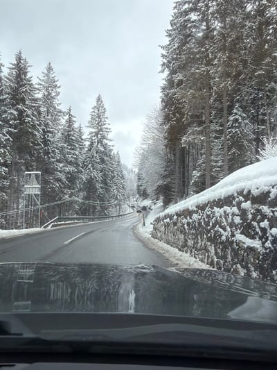 The image displays a winter road scene in Achenkirch, Austria, viewed from inside a vehicle. A two-lane asphalt road, wet and partially cleared of snow, curves gradually to the left and recedes into the distance. White lane markings are visible.

On the left side of the road, a metal guardrail runs alongside the curve, backed by a dense coniferous forest (primarily spruce or fir trees) heavily laden with snow. A utility pole structure with overhead wires is visible near the guardrail in the foreground.

On the right side, a rugged stone retaining wall, partially covered in snow and ice, supports a steep, snow-covered embankment. This slope is also densely covered with snow-laden coniferous trees, with some bare deciduous trees interspersed. Heavy snow blankets the ground and tree branches throughout the scene.

Further down the road, at least two passenger vehicles are visible traveling in the same direction. The sky is overcast. Reflections of icicles or melted snow are visible on the vehicle's hood or dashboard in the lower portion of the image. No people are visible.