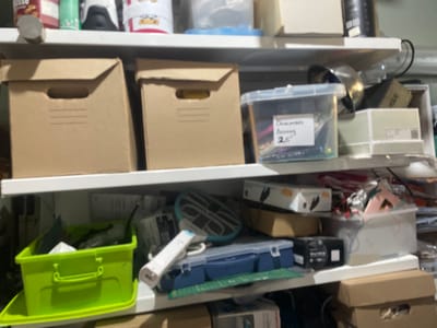 The image shows a cluttered shelf in what appears to be a storage area or a workshop. The shelf is filled with various objects, including cardboard boxes, plastic containers, a green bin, and what looks like a blue toolbox. Some items are organized, while others are jumbled together.

On the top shelf, there are several items that are out of focus, but appear to be food packaging and bottles. Below that, two large cardboard boxes with handles are visible, along with a clear plastic container with some labels. One label has handwritten text that is partially legible, possibly reading "Oncologia" and "25".

The middle shelf contains more diverse items. There is a bright green plastic bin filled with various materials, and a blue toolbox. A white object with a blue tip, resembling a stylus or a pen, is placed next to the toolbox. A circular object with a grid pattern is also present. Further to the right, there are more boxes and containers, some of which are stacked.

The bottom shelf is partially visible and seems to hold more cardboard boxes. The overall impression is one of a functional space where items are stored, possibly for crafting, hobbies, or general household use. There are no people visible in the image. The lighting suggests it is daytime, and the indoor setting provides no clues about the weather or specific location beyond the provided context of Мухавецкі сельскі Савет, Belarus. The scene conveys a sense of practicality and perhaps a need for organization.