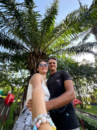In this horizontal, low-angle selfie, a couple is captured against a backdrop of palm trees and lush greenery, suggesting a tropical outdoor setting, possibly Thuận Nam, Vietnam. The sun, partially obscured by the foliage, indicates it's likely daytime. The sky above is clear and blue.

The couple appears to be the primary subjects. On the right, a woman with sunglasses smiles at the camera, her arm extended to take the selfie. She's wearing a white tank top and denim shorts, adorned with a bracelet of various colored beads. On the left, a man with dark hair and a friendly smile is partially visible, wearing a black t-shirt with a faint galaxy print.

They seem to be enjoying a relaxed moment outdoors, possibly on vacation or an outing. The overall mood conveyed is one of happiness and camaraderie. In the background, a stone wall and more trees can be seen, adding to the natural, serene environment. A bright pink flamingo lawn ornament is visible in the lower right portion of the frame, adding a touch of whimsy to the scene. No discernible text is visible in the image.