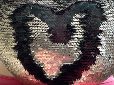 The image shows a close-up view of a pillow or cushion covered in reversible sequins. The sequins are a mix of silver, black, and a pinkish-red hue, arranged to form a pattern that resembles a heart. The pillow is positioned on a pink fabric surface, possibly clothing or another textile. There is no indication of the city of Kamen-na-Obi, Russia, or any other specific location within the image. The lighting is soft, highlighting the texture and shine of the sequins. The scene is static, with no discernible actions or interactions taking place.