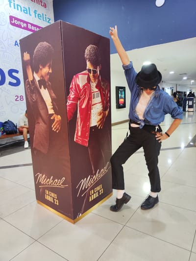 A person dressed as Michael Jackson poses next to a promotional stand for the movie "Michael" in what appears to be a shopping mall or public space. The person is wearing a hat, sunglasses, a blue shirt, and black pants with suspenders. The promotional stand features images of Michael Jackson. The text on the stand is in Spanish, indicating that the movie will be released in cinemas in April 2023. There are people and signs in the background, suggesting that this photo was taken in a public place, likely in the city of Tacna, Peru.