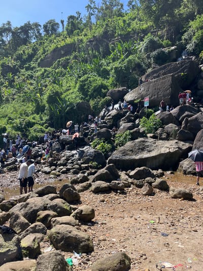 The image depicts a crowded scene at a rocky stream with a waterfall in the background, likely located in Jaflong, Bangladesh. A large number of people are gathered, some are standing on the rocks, others are walking near the water, and a few are sitting. The environment is lush and green, with dense vegetation covering the hilly terrain. The stream is filled with numerous large, gray rocks of various sizes, and the water is shallow in the foreground, appearing somewhat muddy. Several individuals are holding umbrellas, suggesting a sunny day. There is visible litter scattered among the rocks and on the ground, including plastic bags and wrappers. A sign with text in a foreign language is posted on a large rock face. In the foreground, two men are standing in the shallow water, facing away from the camera. The overall impression is a popular tourist or recreational spot, possibly a picnic area, with a natural landscape.