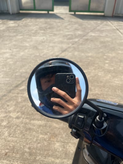 A close-up shot captures a person's reflection in a motorcycle's side mirror. The individual is wearing a black helmet and a blue and red jacket, with a smartphone held in their right hand, angled to take the selfie. The mirror is round and framed in black, mounted on the handle of a black motorcycle. The background, visible in the mirror and beyond, shows a paved outdoor area, possibly a street or driveway, with a light-colored concrete surface and some faint shadows. In the far background, a partially obscured green gate and a light-colored structure are visible. The scene appears to be outdoors, under natural daylight. The location is not specified in the image.