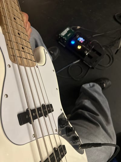 A close-up depicts a white electric bass guitar held by a person wearing grey pants. The guitar is plugged in with a black cable. In the background, slightly out of focus, is a black multi-effects pedal with illuminated buttons and an attached power cord. The setting appears to be indoors, possibly a studio or stage, given the dark flooring.