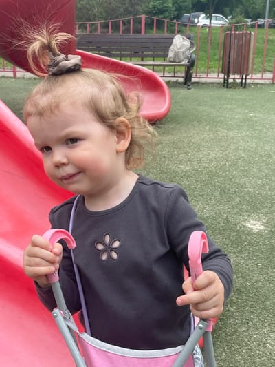 A young girl with blonde hair and a brown hair tie stands beside a pink slide with her left hand holding onto a handle on a pink stroller. The stroller has a pink fabric seat and a gray frame. The girl is wearing a gray long-sleeved shirt with a flower cut-out design. She is smiling at the camera. The scene takes place in a playground in Moscow, Russia, located at coordinates 55.659, 37.591. The playground is characterized by artificial grass, a red slide, and a wooden bench. The playground appears to be empty, with only the girl present. A few trees can be seen in the background, along with a parked white car. The image seems to have been captured on a sunny day.