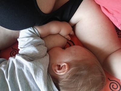 This image captures an intimate overhead view of a mother and her baby, likely indoors. The main subjects are the upper torso and arm of an adult woman, partially clad in a black top or bra, and a baby lying next to her, feeding.

The baby, with light-colored hair and wearing a grey long-sleeved shirt, is positioned with its head against the mother's breast, engaged in breastfeeding. One of the baby's small, chubby hands rests on the mother's chest or arm near the feeding area. The mother's skin appears fair, and a small mole is visible on her upper arm.

They are resting on an orange surface, which appears to be bedding or a blanket, featuring a repeating black spiral or swirl pattern. A pink fabric, possibly another blanket or piece of clothing, is visible on the right side of the frame, contrasting with the orange. The lighting suggests daytime, with soft, natural light illuminating the scene. The overall atmosphere conveys a sense of quiet nourishment and closeness between the mother and child.

There is no discernible text visible in the image. The location context provided is Teykovo, Russia.