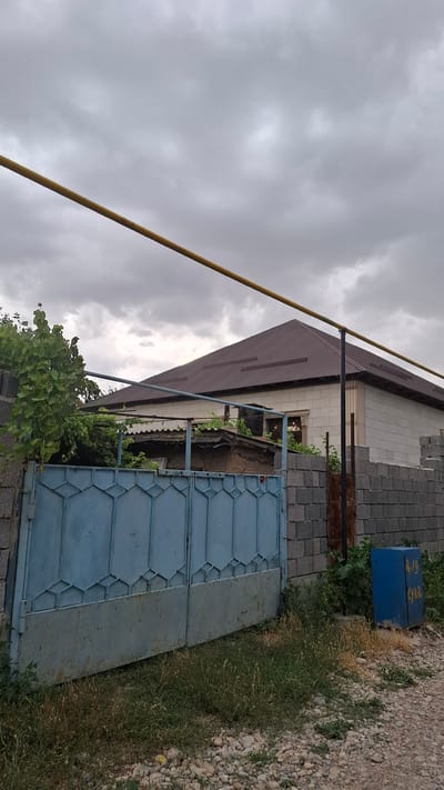 The image depicts the exterior of a residential property in Шұбарсу, Kazakhstan. A blue, corrugated metal gate with a decorative geometric pattern is partially open on the left side of the frame. Behind the gate, a low-slung, weathered structure with a corrugated roof is visible, overgrown with green foliage. To the right of the gate, a brick wall forms the boundary of the property. Beyond the gate and the foreground structure, a larger house with a dark, sloped roof made of corrugated metal is visible. This house has walls constructed of light-colored concrete blocks with some decorative detailing. A yellow pipe runs horizontally across the upper portion of the image. In the foreground, the ground is covered with gravel and patches of dry grass and weeds. To the far right, a blue electrical box with yellow markings, possibly numbers and text, is situated near the brick wall. The sky is overcast with heavy, gray clouds.