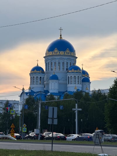 This image captures a white and blue Orthodox church with golden ornamentation, featuring multiple domes topped with crosses. The church is situated near a road with several cars in traffic. The sky is a light gray and orange, indicating either sunrise or sunset. Lush green trees line the background, suggesting a natural setting within an urban or suburban area. Several road signs are visible, including traffic signals, speed limit signs (one indicating 40), and directional signs. One sign appears to read "ул. ОСТРОВСКОГО" which translates to "Ostrovskogo Street". The overall scene suggests a quiet moment during a transitional time of day, possibly dusk or dawn, with the traffic indicating normal daily activity. The lighting is soft, contributing to a serene atmosphere.