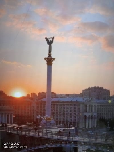 The image shows a sunset over a city with a tall monument in the foreground. The sky is filled with pink and orange clouds, and the sun is low on the horizon, casting a warm glow over the scene. The monument appears to be an angel with outstretched wings, standing on a column. In the background, there are several buildings, some of which are illuminated. A bridge with people walking on it is visible in the lower part of the image. The timestamp "OPPO A17 2026/07/26 22:01" is present in the lower left corner. The location is identified as Qumozek, Kazakhstan.