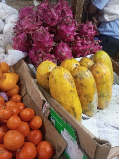 The scene depicts a vibrant fruit stall or market display in Lê Thanh Nghị Ward, Vietnam. In the upper-center, a substantial pile of approximately thirty-five ripe, magenta-colored dragon fruit (pitaya) is arranged. To the mid-right, a row of eight elongated, yellow papayas, exhibiting some green discoloration and dark blemishes, rests on a light-colored surface, possibly paper, within a cardboard container. In the lower-left, a collection of numerous small, round, bright orange mandarins or tangerines is visible, packed within a brown cardboard box. Some mandarins retain small green stems. White protective foam netting is partially visible to the left of the dragon fruit. A green-labeled carton is visible beneath the papayas. In the blurred background, upper-right, a person wearing a light blue shirt is partially visible, facing away from the viewer. The overall environment suggests an indoor or covered market setting, with visible wooden structures in the background.