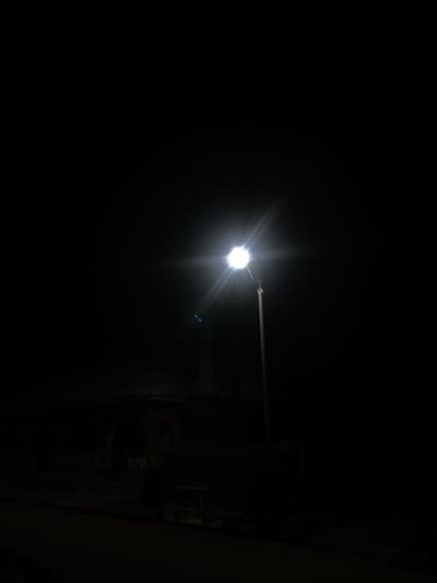 This nighttime image, captured in Aigiere, Nigeria, displays a solitary, intensely bright street light positioned slightly right of center on a vertical pole. The light source generates a pronounced starburst effect and distinct blue lens flares. The overwhelming majority of the scene is enveloped in deep darkness, characteristic of minimal ambient light. In the mid-ground, to the left of the illuminated pole, faint, silhouetted architectural outlines are visible, suggesting a building structure with a prominent roofline or dome. The immediate foreground consists of a dark, undifferentiated surface, likely a road or path. No individuals, discernible actions, or other significant objects are identifiable within the frame.