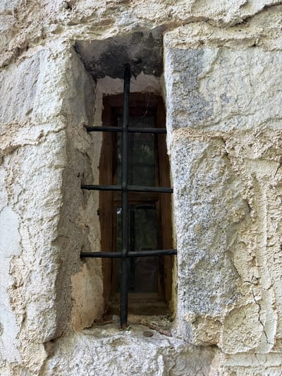 A barred window set into a rough stone wall is located in Govedartsi, Bulgaria. The window frame is made of wood and contains panes of glass. A grid of black metal bars covers the exterior opening. The stone wall appears old and weathered with textured surfaces. The light suggests it is daytime. No people are present.