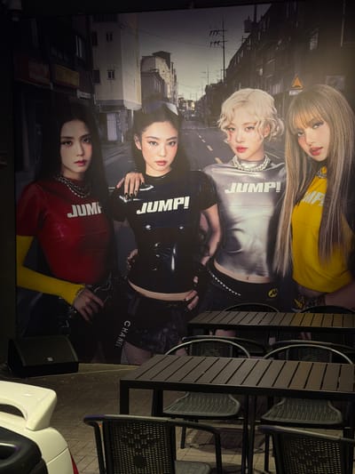 The image shows a vertically oriented poster featuring four women, all positioned against an urban backdrop of buildings. The women are dressed in garments with the word "JUMP!" prominently displayed. A table and chairs are situated near the poster. The location is not explicitly stated but is in the city of Linghe, China.