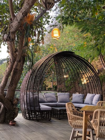 This media file captures an outdoor seating area, likely a lounge or restaurant patio, set in a lush, tropical environment. The focal point is a large, woven, dome-shaped structure resembling a bird's nest, enclosing a comfortable sectional sofa with light gray cushions. Adjacent to the dome, a small coffee table made of woven material sits in front of the sofa. To the right, a wooden table is paired with two wicker chairs, suggesting a dining or gathering space.
The setting is characterized by abundant greenery, with large trees and dense foliage surrounding the seating area. A prominent tree with a thick, textured trunk stands to the left, adorned with epiphytic plants, possibly staghorn ferns, and hanging roots. A decorative, woven pendant lamp with a warm, glowing light hangs from a tree branch above, casting a soft illumination.
The scene appears to be set during the late afternoon or early evening, as suggested by the warm, diffused light filtering through the trees and the glowing lamp. The sky visible in the background is a soft, hazy white, indicating daylight. The overall mood is tranquil and inviting, emphasizing relaxation and connection with nature.
The wooden deck flooring on which the furniture rests suggests a raised platform or an elevated seating area, possibly overlooking a natural landscape. A small, reddish-brown clay pot is visible on the deck near the base of the large tree. No people are visible in the image. There is no visible text in the media file. The context provided indicates this location is in Labuan Bajo, Indonesia, which aligns with the tropical flora and outdoor ambiance.