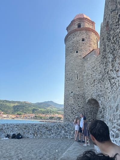 In the city of Collioure, France, a stone tower with a pink dome stands prominently against a clear blue sky. The tower, part of a larger stone fortification, is textured with various sizes of grey stones and has small square windows. To the right of the tower, a section of the stone wall extends outward, revealing an arched entrance. In front of the wall, a group of people are visible. A man in a white shirt and dark shorts stands with his back to the camera, facing a young woman in a light-colored top and skirt, and another young woman in a striped dress. They appear to be looking out towards the water.

In the background, the Mediterranean Sea is visible, with a stone seawall bordering it. Beyond the sea, the town of Collioure stretches along the coast, featuring buildings with red-tiled roofs and the green, hilly landscape of the Pyrenees Mountains rising behind it. The ground in the foreground is a sandy or gravelly beach. Several backpacks and bags are placed on the ground near the seawall. The scene captures a view from a historic site overlooking the coastal town and its natural surroundings.