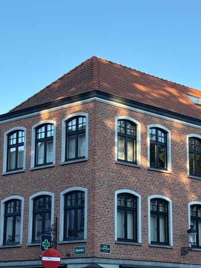 The image shows a corner of a brick building with several arched windows. The building is located in Bruges, Belgium, as indicated by the street sign visible in the lower right portion of the image which reads "SMIDSTRAAT". The street sign in the lower left portion of the image is a red circle with a white horizontal bar, indicating "no entry". A green shield with a white cross is also visible. The sky is clear and blue, with the sun casting shadows on the building's roof.