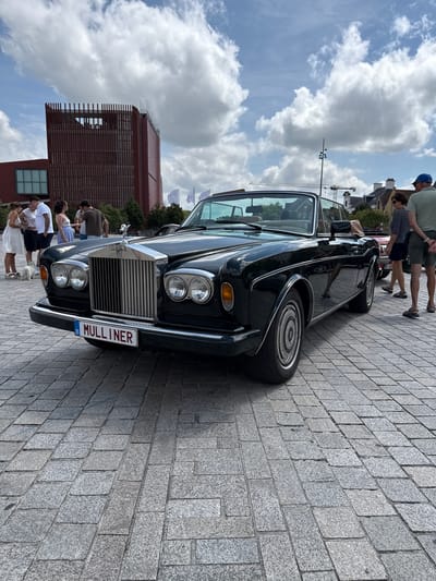 In Bruges, Belgium, a dark green Rolls-Royce Corniche convertible is parked on a cobblestone square. The car's front is prominently displayed, featuring its iconic grille, dual headlights, and a license plate that reads "B MULLINER". Several people are gathered around the car, observing it. In the background, a modern red building with vertical slatted detailing stands out against a sky filled with white, puffy clouds. The scene appears to be an outdoor event or exhibition, possibly a car show.