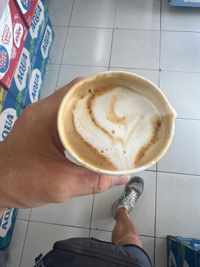 A person is holding a cup of coffee with latte art in their hand. The person is wearing sneakers and a backpack. Stacks of bottled water, labeled "AQUA," are visible near the cup. The scene is likely in Celuk, Indonesia. The background features a tiled floor and walls.