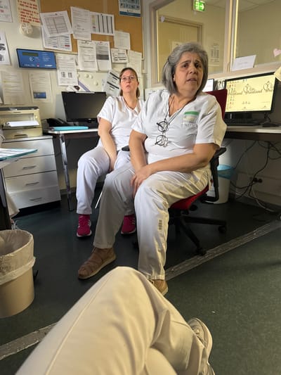 The image depicts an interior scene, likely an administrative or staff area within a healthcare facility in Tourcoing, France. Three individuals are present. In the foreground, the lower legs of a person wearing light-colored trousers and sneakers are visible, with ankles crossed. In the mid-ground, a woman with gray hair and glasses hanging from her white scrub top is seated in a red office chair, looking upwards with an open-mouthed expression. She wears light brown sandals. Behind her and slightly to the left, another woman with brown hair and glasses, also in white scrubs, is seated, looking upwards with a neutral expression. She wears bright pink athletic shoes.
The environment includes a cork bulletin board covering the back wall, densely affixed with various printed documents and notices. Below the board, two desks are visible. The left desk holds a Toshiba printer, a dark computer monitor, and a blue folder, with a light-colored filing cabinet underneath. A white waste bin is on the floor to the far left. The right desk features a computer monitor displaying a light-colored screen with "Droland Dro" partially visible text, with power cables underneath. The floor is dark green, possibly linoleum, with a worn strip of white tape or sealant running horizontally. A light-colored door with a green "Exit" sign is visible in the upper right background.