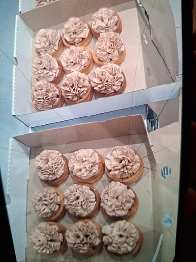 The image shows two open cardboard boxes filled with small tarts. The tarts have a light brown crust and are topped with a swirl of light brown frosting, resembling a flower. The boxes appear to be arranged on a surface, with the top box slightly elevated above the bottom box. The background is out of focus and appears to be a light blue. There are several thin, dark lines across the image, possibly scratches on a screen or lens. The location is not identifiable from the image.