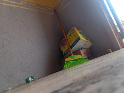 The image depicts an outdoor scene in Barakin Angundi, Nigeria, likely in a back alley or storage area. A green broom with a yellow handle rests against a textured gray wall, leaning against stacked cardboard boxes. One prominent yellow box features an image of a chicken and text indicating "REAL CHICKEN SOURCE" and "INSTANT NOODLE." Adjacent to it is a smaller box labeled "Kremela." In the lower left corner of the image, a small green and white package is visible on the ground, along with some scattered debris. The ground appears to be made of concrete or a similar material. The lighting suggests it is daytime, with sunlight entering from the right side of the frame, creating strong shadows and highlights. The overall environment appears somewhat weathered and utilitarian.