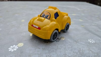 A yellow toy car with cartoonish eyes and an open mouth with white teeth is positioned on a table covered with a patterned cloth. The cloth has a light gray base with a repeating pattern of white and gold flowers. The car has black wheels with gray rims. The background is blurred, suggesting a shallow depth of field, and appears to be an indoor or covered outdoor setting. There is no indication that this image is related to Samarkand, Uzbekistan.