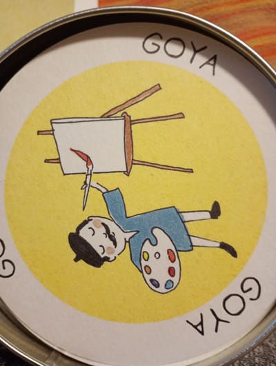 The image is a close-up shot of a round, yellow surface with cartoon drawings. At the top, the word "GOYA" is written in black, stylized letters. In the center, a cartoon of an artist is depicted. The artist is wearing a blue smock and holding a palette with colorful paint blobs. They are standing on their head and painting on an easel, with a red streak of paint on the canvas. To the left of the artist, the letter "G" is visible, partially cut off by the image border. At the bottom right, the word "GOYA" is written again, upside down relative to the top text. The entire drawing is contained within a silver-colored rim, suggesting it's a lid of some sort, possibly a tin or a box. The background outside the yellow circle appears to be a textured surface, possibly fabric, with horizontal stripes of orange and yellow. This image does not contain any identifiable elements or landmarks that would allow for its location to be determined in the city of Rosenheim, Germany.