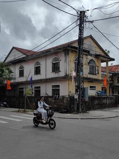 The image displays an urban street scene in Phú Xuân Ward, Vietnam, under a heavily overcast sky. A female, wearing a white long-sleeved shirt, white pants, and a dark backpack, rides a dark-colored electric bicycle equipped with a front basket. She is positioned facing slightly left, gripping the handlebars, and navigating from the left side of the frame toward the right across an asphalt street. A prominent white pedestrian crossing is marked on the asphalt in the foreground.

In the background, a two-story building occupies the corner of an intersection. The building's left section is painted a faded light pinkish-white, while the right section is a degraded yellow, both exhibiting extensive dirt and weathering. It features a red-tiled hip roof. The upper story contains multiple arched windows with dark frames, some appearing open. The lower story has rectangular windows. A dark, stained concrete wall serves as the building's perimeter fence. Attached to this wall are a yellow informational sign with graphics on the left and a white sign further to the right. An orange rectangular flag is visible on the left section of the wall, and a blue rectangular flag is near the center.

A tall, dark gray metal lattice utility pole stands directly at the building's corner, heavily laden with numerous crisscrossing black electrical and communication wires. A gray utility box is mounted on the lower part of this pole. A pink rectangular flag hangs near the utility pole, and another orange flag is visible further to the right, adjacent to an unseen structure. A young tree supported by wooden stakes is positioned against the concrete wall on the left. A parked scooter is partially visible behind the wall on the far left of the frame.