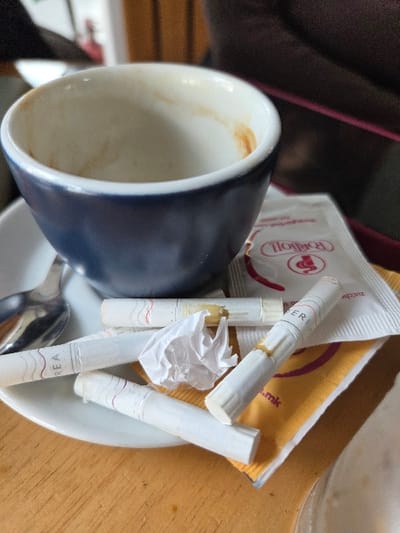 The scene depicts a close-up of a table surface within an establishment located in Gevgelija, North Macedonia. A dark blue ceramic coffee cup, its white interior stained with dried brown residue from a consumed beverage, rests on a white saucer. A silver-colored spoon is positioned on the left side of the saucer. Scattered across the saucer and adjacent to the cup are four white cigarette filters; one filter bears the partial text "REA", another shows "TER", and several exhibit yellowish-brown tar discoloration. A small, crumpled piece of white paper or wrapper is situated among the filters. Beneath some of these items, a white sugar packet labeled "ROQIOLL" is visible, along with a partially obscured yellow-orange sugar packet displaying a '.mk' internet domain, supporting the North Macedonian context. The table surface is light brown wood with discernible grain. The background is out of focus, showing indistinct dark and reddish elements consistent with an indoor setting.