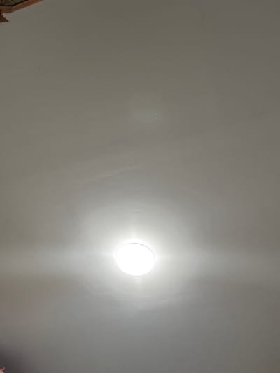 The image is a shot looking directly up at a white ceiling with a recessed, circular light fixture. The light fixture is on and emitting a bright white light. The rest of the ceiling is a uniform off-white or light gray color. The light and the surrounding ceiling seem to be the only identifiable elements. The location is not evident from the image.